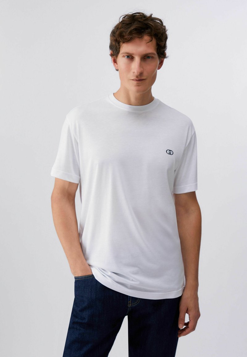Homme portant un t-shirt blanc uni à manches courtes avec un petit logo noir sur la poitrine, jean bleu foncé, debout sur un fond clair neutre.