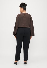 Braunetransparente Bluse mit Puffärmeln, Rückenschlitzdetail, kombiniert mit schwarzen maßgeschneiderten Hosen und schwarzen High Heels.