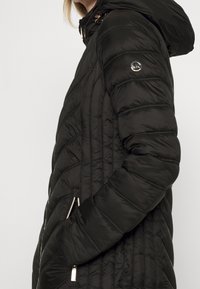 Schwarze gesteppte Pufferjacke mit Kapuze, versehen mit diagonalen Nähten, silbernen Reißverschluss-Taschen und einem runden Logo-Akzent am Ärmel.