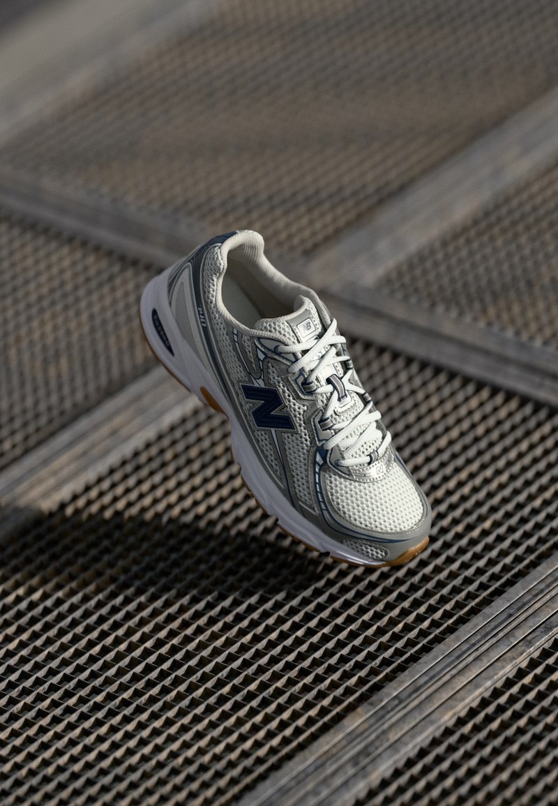 Chaussure de sport New Balance blanche et grise avec logo bleu marine, flottant au-dessus d'un fond à grille métallique texturée.