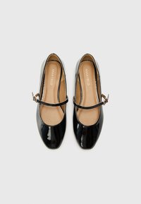 Black patent leather Mary Jane flats with a rounded toe, an ankle strap with a buckle, and a smooth tan insole.