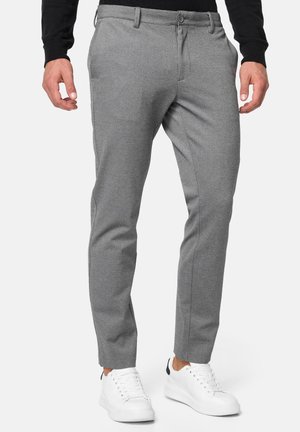 Homme portant un pantalon gris slim et des baskets blanches, debout les mains détendues sur les côtés sur un fond uni.