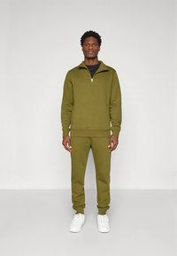 Sudadera con cremallera en verde oliva y joggers a juego, hechos de tela suave. Cuenta con un cuello de media cremallera y puños acanalados. Combinado con zapatillas blancas.