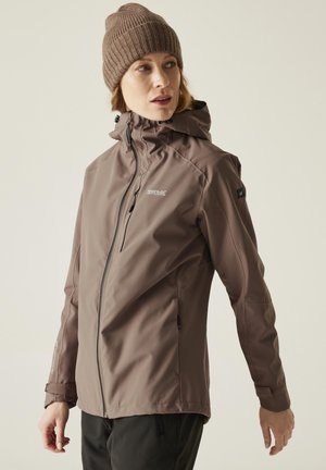 Donna con berretto marrone e giacca outdoor con zip e cappuccio, che guarda di lato su uno sfondo semplice.