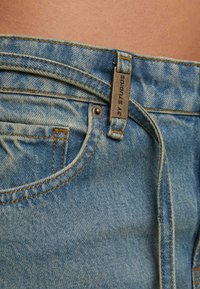 Kadar izbliza remena plave traperice i stražnjeg džepa s metalnom oznakom na kojoj piše "2Y STUDIOS" na golom struku.