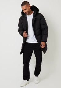Czarna kurtka puchowa z kapturem wykończonym futrem, zapinana na zamek z przodu, z bocznymi kieszeniami, połączona z czarnymi spodniami i białymi trampkami.