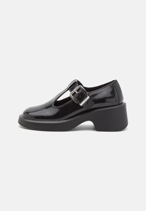 Zapato de charol negro con diseño de tira en T, hebilla ajustable, punta redonda y tacón grueso. Textura lisa con detalles mínimos.