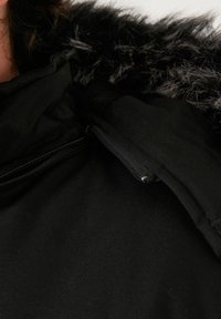 Čierna bunda s umelým kožušinovým golierom, s uzáverom na zips a textúrovanou tkaninou. Kapucňa je tiež podšitá mäkkým materiálom.