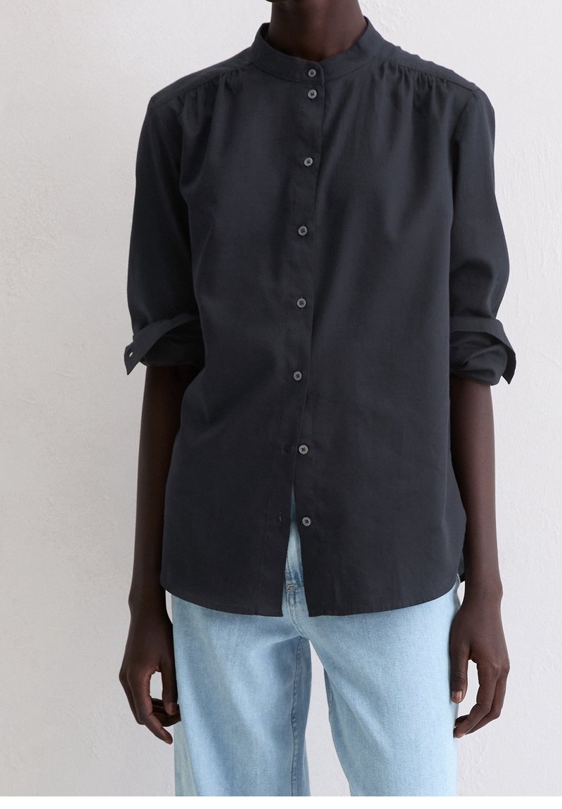 Personne portant une chemise noire à manches longues boutonnée avec les manches retroussées et un jean bleu clair, debout devant un fond blanc uni.