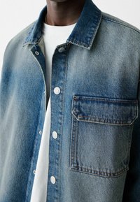 Camicia in denim in varie tonalità di blu, con chiusura a bottoni, due tasche e cuciture a contrasto. Texture liscia e design casual.