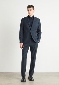 Abito blu navy con giacca monopetto e pantaloni con frontale piatto. Il tessuto appare liscio; include un collo a cratere e chiusura a bottoni. Scarpe nere.
