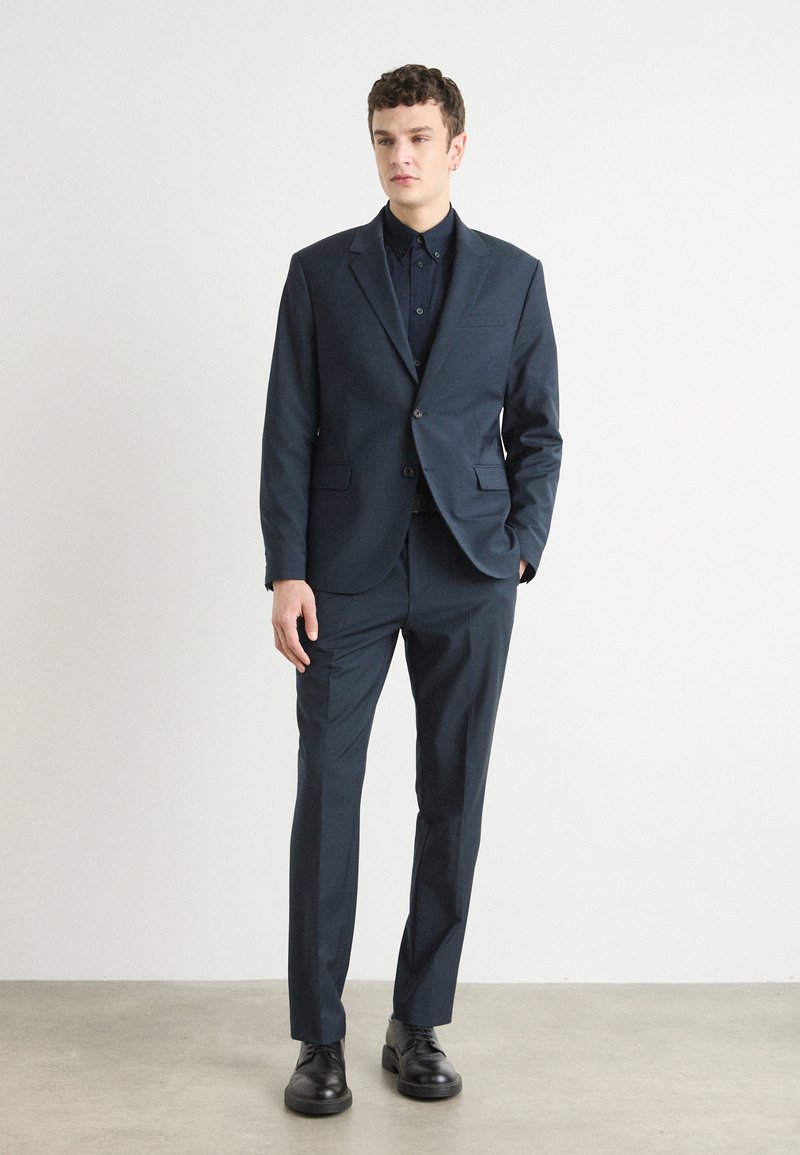Costume marine avec un blazer à simple boutonnage et un pantalon à taille plate. Le tissu semble lisse ; comprend un revers cranté et une fermeture à bouton. Chaussures noires.