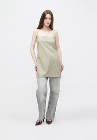 Khakifarbenes ärmelloses Kleid mit quadratischem Ausschnitt, kombiniert mit hellgrauen, ausgestellten Jeans und burgunderroten, spitz zulaufenden Schuhen. Glatte Stofftextur.