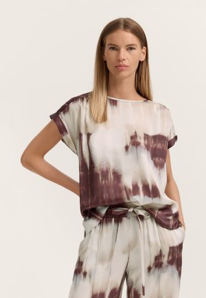 Frau mit glatten blonden Haaren, die ein locker sitzendes kurzärmeliges Batikshirt und passende Kordelzughosen in Creme- und Brauntönen trägt.