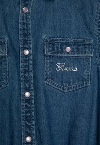 Denim shirt in dark blue with pearl snap buttons. Features two chest pockets, one adorned with rhinestone accents spelling "Guess."