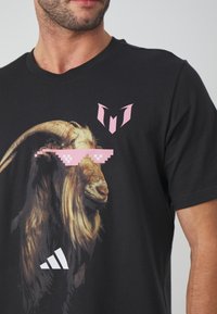 Musta puuvillapaita, jossa on graafinen kuva vuohesta, joka käyttää pikselöityjä vaaleanpunaisia aurinkolaseja, sekä valkoinen adidas-logo edessä. Lyhyet hihat, pyöreä kaula-aukko.