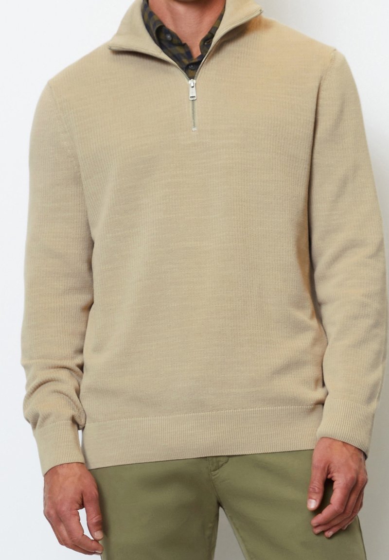 Homme portant un pull beige à fermeture éclair jusqu'au quart, par-dessus une chemise à carreaux foncés, avec un pantalon vert, debout devant un fond uni.