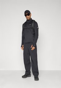 Man in een zwart outdoor outfit met een pet, zonnebril, rugzak, lange mouw rits-top, cargobroek en wandelschoenen staan tegen een witte achtergrond.