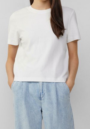 Mujer con una camiseta blanca lisa de manga corta, ligeramente metida en vaqueros sueltos de mezclilla azul claro, con las manos en los bolsillos.