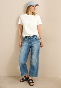 Witte T-shirt met korte mouwen en geborduurde details, lichtblauwe denim wijde jeans en donkerblauwe sandalen. Pet in bijpassend lichtblauw.