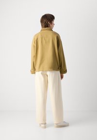 Khaki cotton jacket with a slightly oversized fit, featuring a ribbed collar and buttoned cuff accents, paired with off-white trousers.