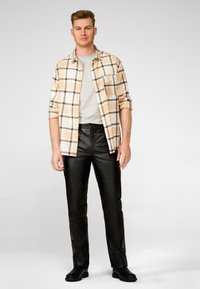 Herenoutfit met een beige en geruite overhemden, grijze t-shirt en zwarte leren broek. Het overhemd heeft lange mouwen en een zak.