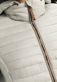 Polstret jakke i lys grå med et elegant design. Har en kontrastfarvet brun og sort lynlås samt vandret quiltet tekstur.