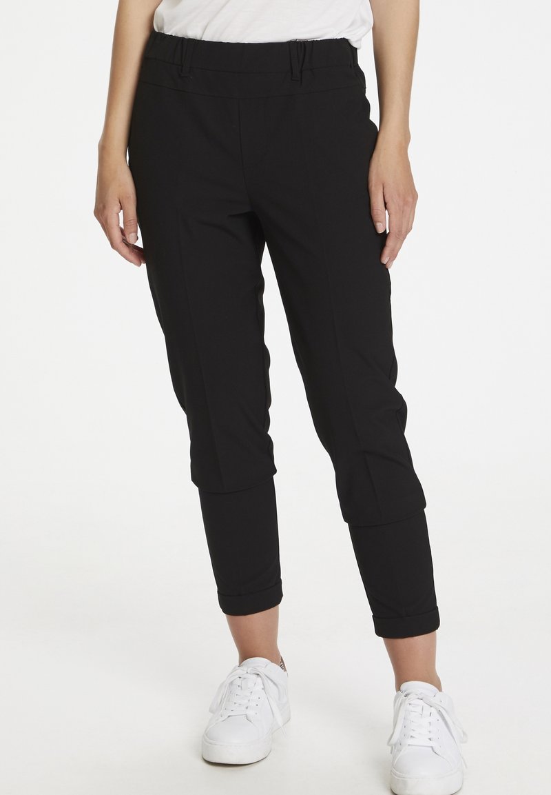 Pantalons de jogging noirs, ajustés avec une taille élastique et des poignets roulés, fabriqués dans un matériau extensible. Associés à des baskets blanches.