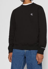 Homme portant un sweat-shirt noir Calvin Klein avec un petit logo blanc cK sur la poitrine et le texte Calvin Klein sur le bord de la manche gauche, associé à un jean bleu.