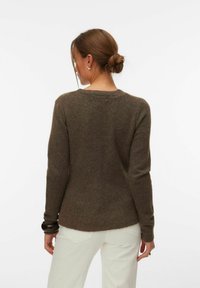 Frau mit braunen Haaren in einem Dutt, die einen braunen Langarmpullover und weiße Hosen trägt, mit dem Rücken zur Kamera vor einem einfarbigen weißen Hintergrund.