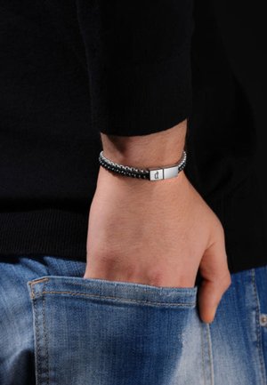 Bracciale intrecciato nero con chiusura in tonalità argento, caratterizzato da un accento metallico rettangolare. Indossato su un polso, abbinato a un denim.
