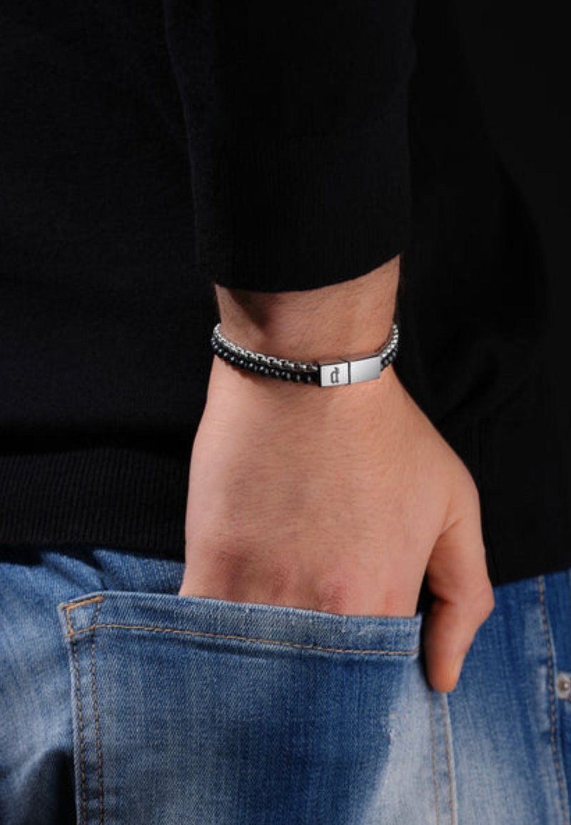 Black braided bracelet with silver-tone clasp, featuring a rectangular metal accent. Worn on a wrist, paired with denim.