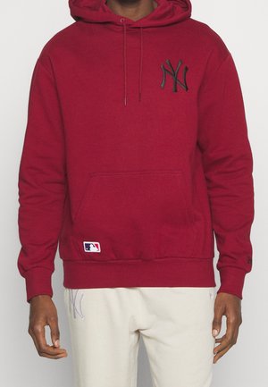 Persona que lleva una sudadera con capucha roja con el logo negro de los New York Yankees y pantalones blancos con un logo gris claro en el muslo izquierdo.