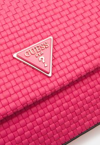 Rosa vävd handväska från Guess med en triangulär guldfärgad logotyp och kontrasterande sömmar. Mjuk textur med en strukturerad design.