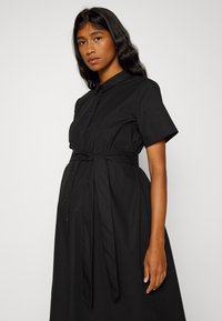 Vestido preto com botões, mangas curtas e colarinho. Apresenta uma faixa na cintura e uma saia fluida. Feito de um tecido suave com uma textura nítida.