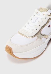 Białe trampki z złotymi akcentami, wykonane z zamszu i skóry, z płaskimi białymi sznurowadłami oraz gumową podeszwą. Na boku znajduje się logo marki.