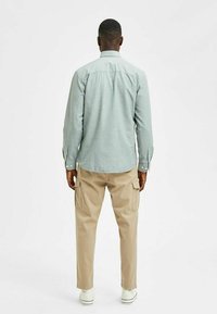 Lichtblauw button-up shirt met lange mouwen, rugpand en contrasterende knopen, gecombineerd met beige cargobroek en witte sneakers.