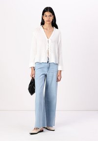 Bluză albă texturată cu închidere prin legare, combinată cu blugi largi de culoare albastru deschis și balerini bej. Ținând o geantă neagră. Outfit simplu și casual.