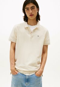 Beige Poloshirt mit weicher Textur, kurzen Ärmeln und einem Kragen mit zwei Knöpfen. Verfügt über ein kleines Logo auf der Brust. Trägt man mit blauer Jeans.