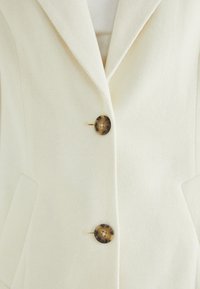Blazer avorio in tessuto testurizzato, con colletto a punta e due bottoni marroni decorati davanti. Include due tasche laterali.