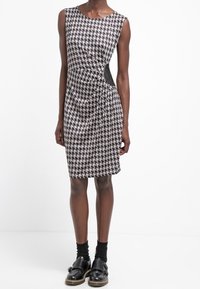 Šaty s černobílým vzorem houndstooth, fitted siluetou a bočním výřezem, doplněné černými ponožkami a mokasínami.