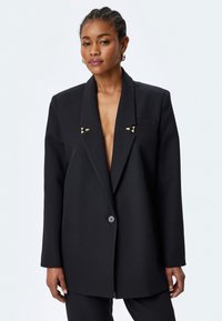 Zwarte oversized blazer gemaakt van een gestructureerde stof, met een diepe V-hals, twee gouden accentclips en een enkele knoopsluiting.