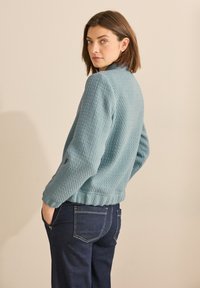 Giacca blu texture con colletto alto, polsini a coste e orlo accorciato, abbinata a pantaloni di jeans scuri.
