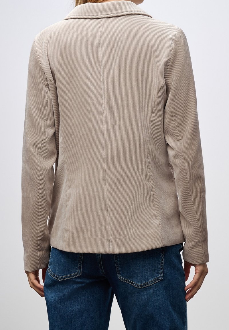 Personne portant une veste beige en velours côtelé et un jean en denim bleu, vue de dos sur un fond clair uni.