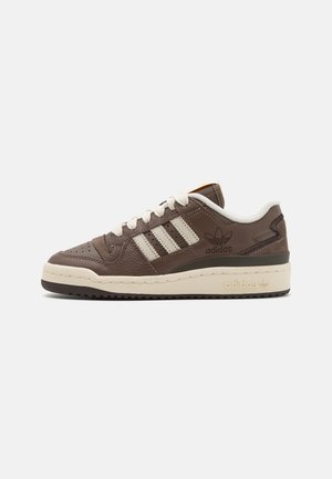 Baskets basses Adidas marron avec des rayures blanches, des lacets blancs et une semelle beige sur un fond gris clair uni.