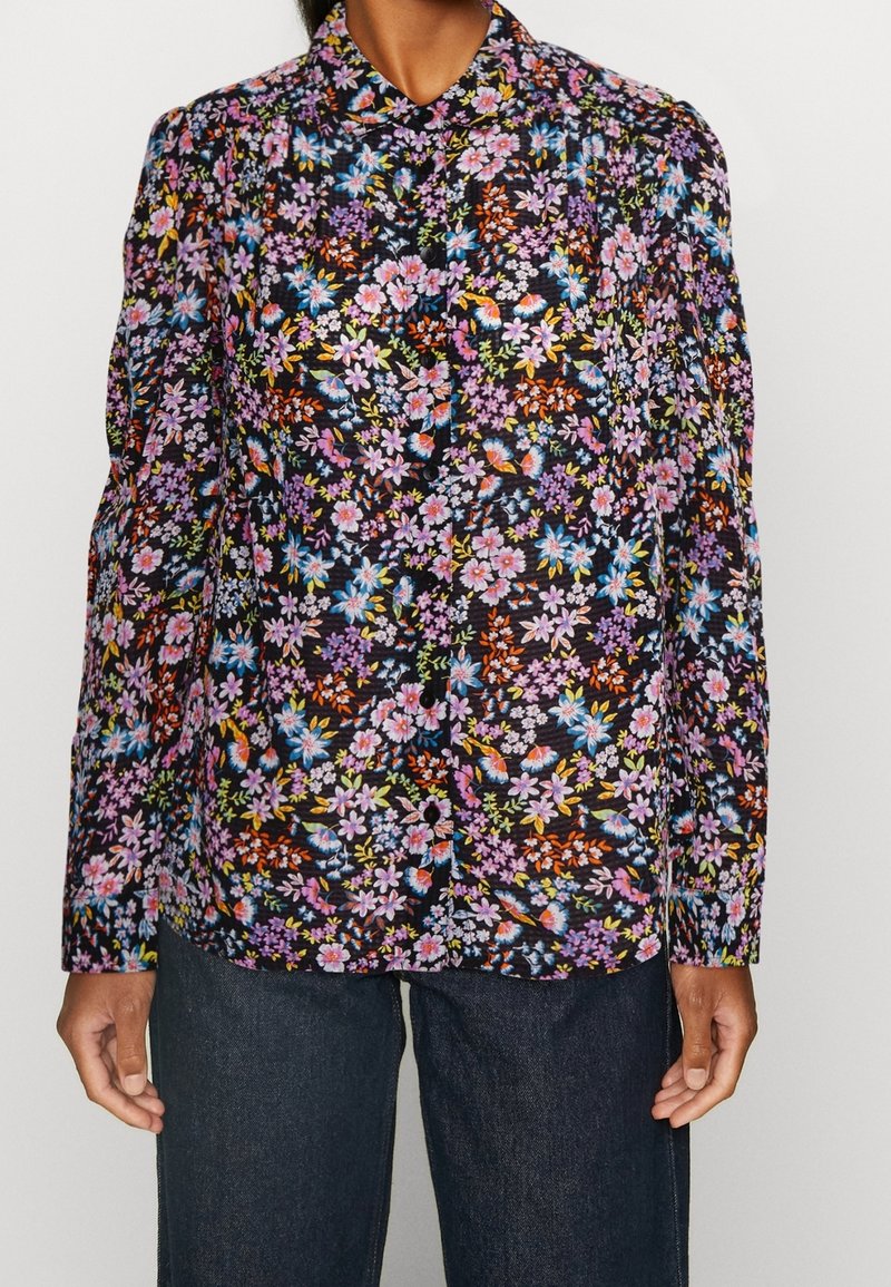 Blouse à manches longues avec un fond noir et un motif floral multicolore dense, portée avec un jean bleu foncé.