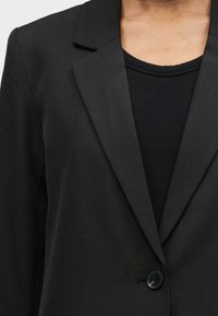 Μαύρο σακάκι με ένα κουμπί και πέτο με σχισμές, φορεμένο πάνω από μαύρο ριμπ μπλουζάκι με στρογγυλή λαιμόκοψη, που αφήνει ακάλυπτη την περιοχή του πάνω μέρους του στήθους και των ώμων.