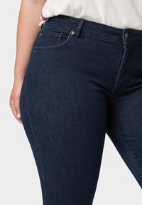 Pantalones vaqueros de mezclilla elásticos de color azul oscuro con una textura suave, que cuentan con cinco bolsillos y detalles de metal plateado. Diseño de cintura de altura media.
