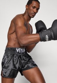 Calções de boxe em cetim preto com fendas laterais, cós elástico e logótipo branco "Venum". O boxeador usa luvas pretas, exibindo os músculos dos braços.