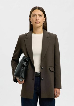 Vrouw met stijlbruin haar, zilveren oorringen, een bruine blazer, wit topje, donkere jeans en een zwarte leren handtas in haar hand.